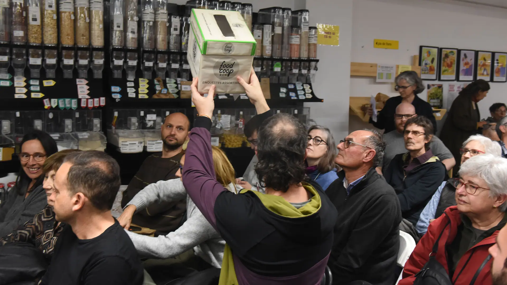 A l’assemblea, una persona aixeca una urna de cartró de FoodCoop BCN per sobre de la gent asseguda, amb els dispensadors de granel al fons.