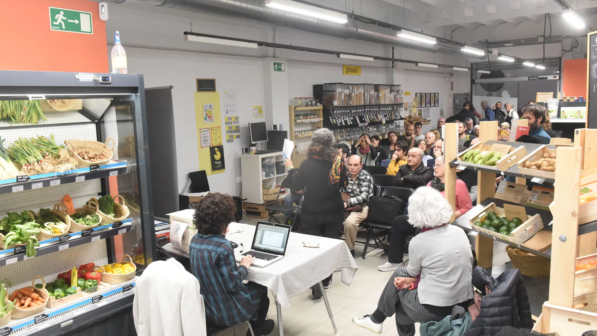 Assemblea de sòcies dins del súper: gent asseguda entre una nevera de verdura i les prestatgeries de granel, i una persona amb un portàtil en una taula.