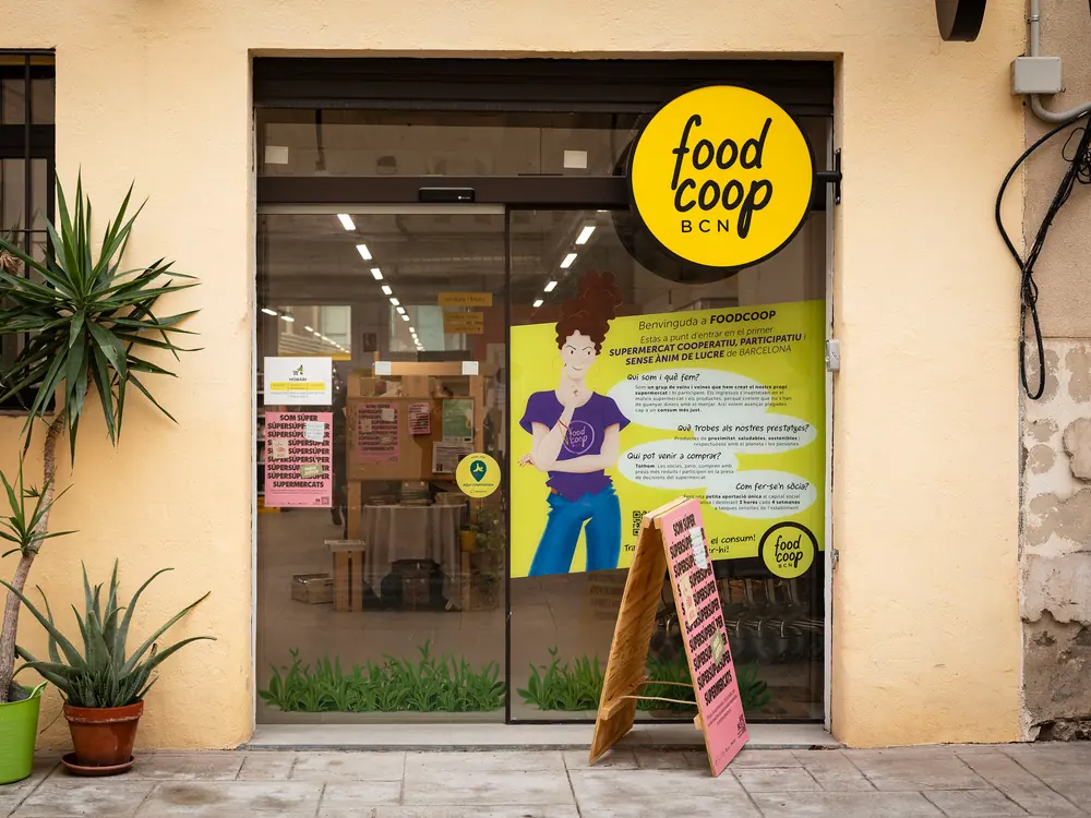 Façana del súper: parets ocre, el rètol rodó groc de FoodCoop BCN, la porta de vidre amb un cartell il·lustrat i un cavallet al carrer.