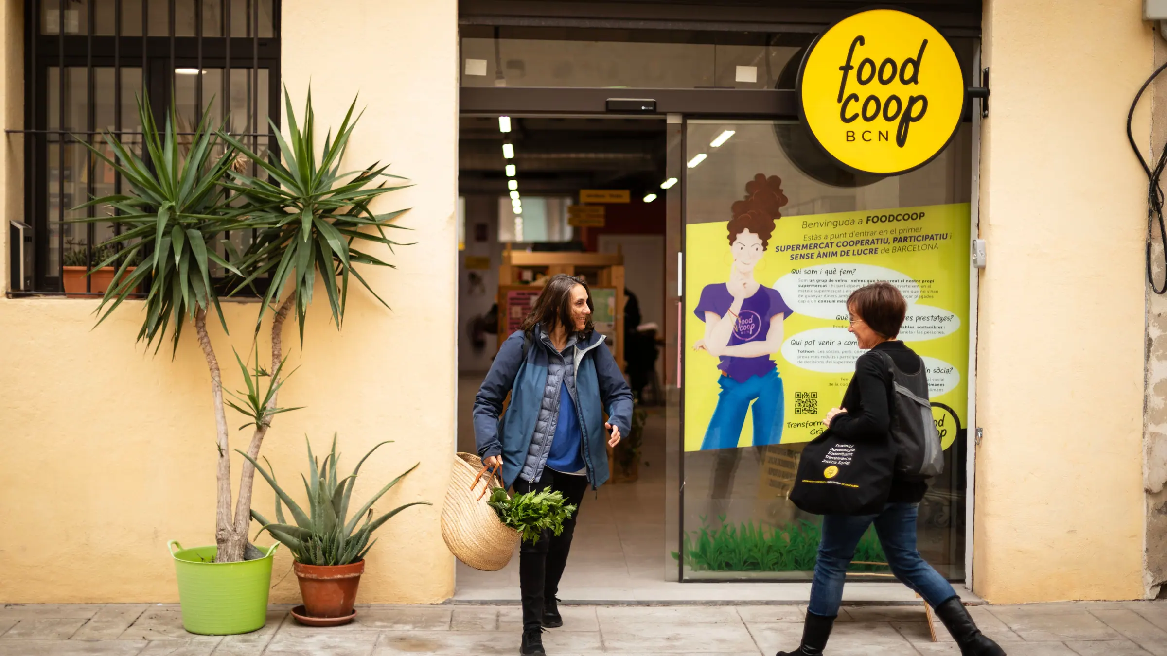 Dues sòcies surten del súper sota el rètol rodó groc de FoodCoop BCN. Una porta un cabàs de vímet amb verdura.
