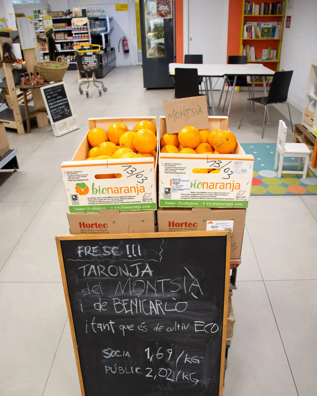 Una pissarra escrita amb guix davant de dues caixes de taronges: «Fresc!!! Taronja del Montsià i de Benicarló, i tant que és de cultiu eco».