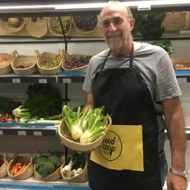 El Jaume, amb el davantal groc i negre de FoodCoop BCN, sosté una panera de fonolls davant dels prestatges de fruita i verdura.