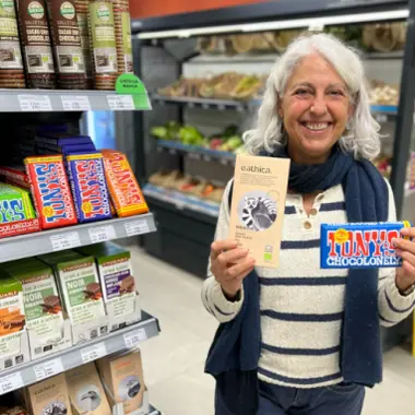 La Rosa, amb cabells blancs i un jersei de ratlles, somriu amb una rajola de xocolata a cada mà, una d’eathica i una de Tony’s Chocolonely, al costat del prestatge de les xocolates.