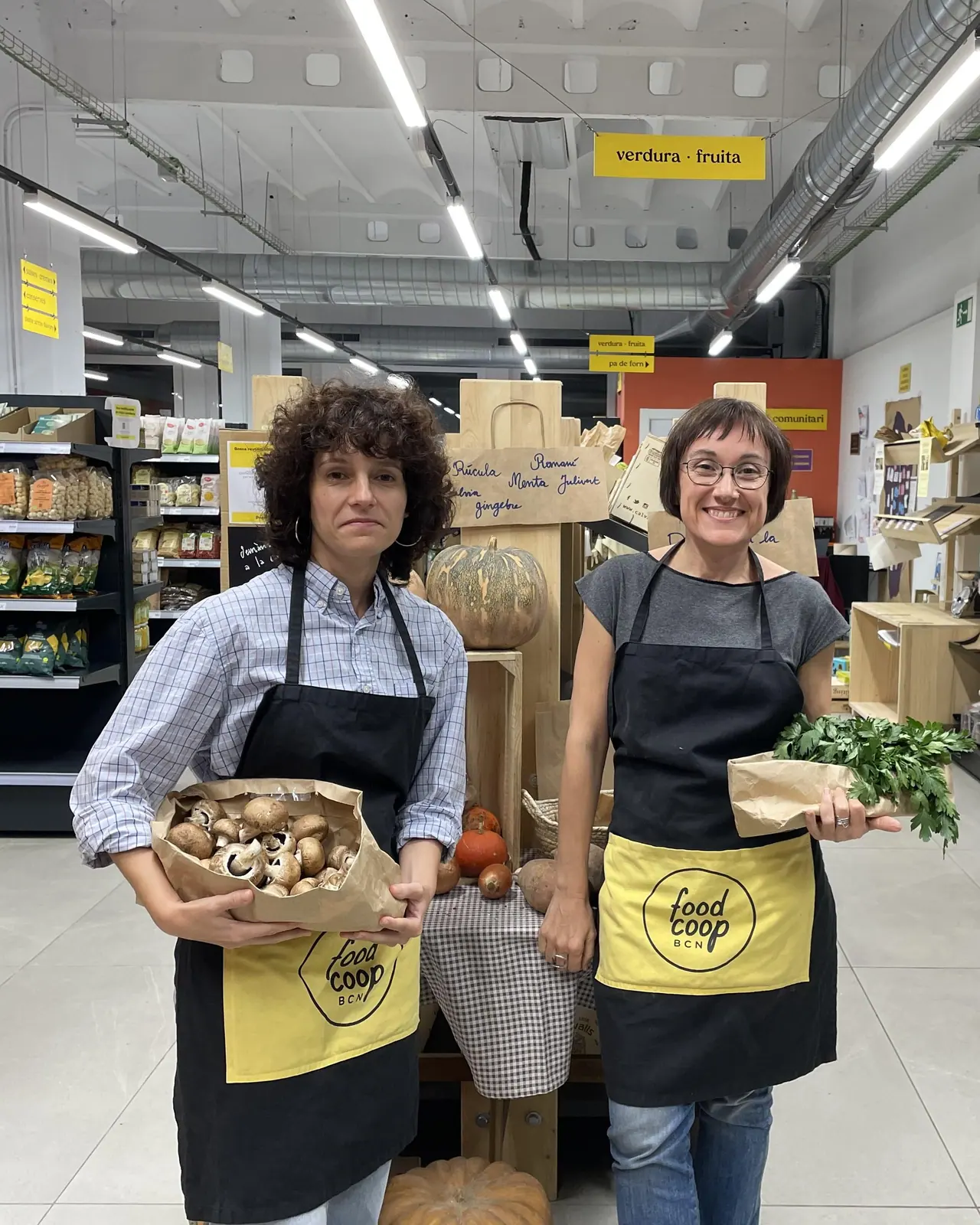 Dues sòcies amb el davantal groc i negre de FoodCoop BCN somriuen entre caixes de verdura: una sosté un paquet de bolets i l’altra un manat de julivert.