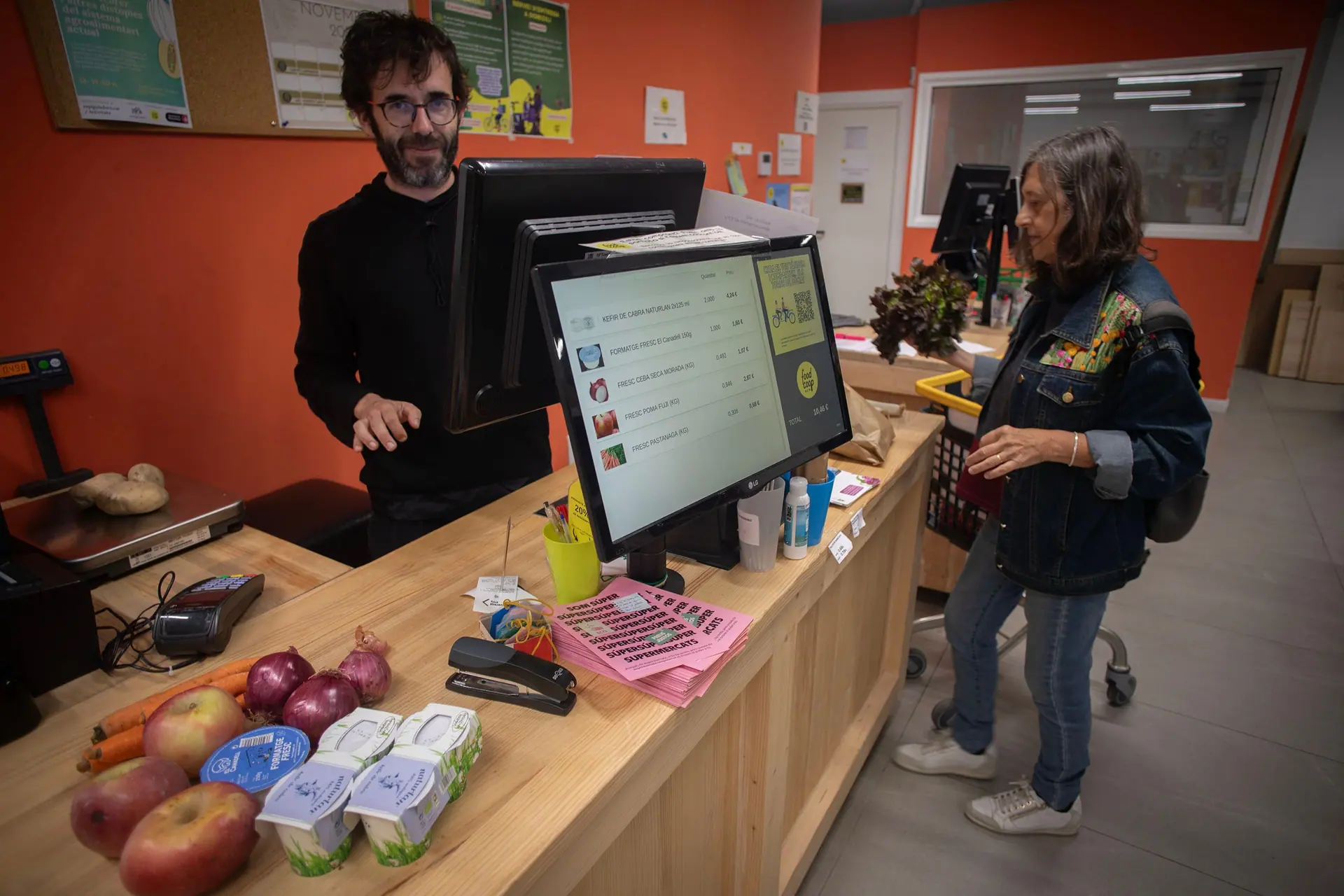 Un torn a caixa: un home cobra davant la pantalla amb la llista de productes i una persona espera amb un carro groc. Al taulell hi ha pomes, cebes i iogurts.