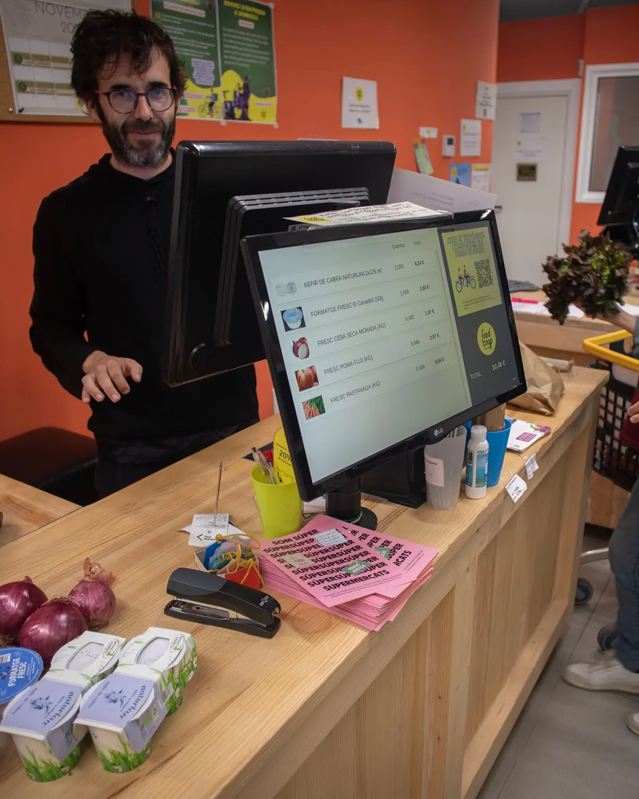 Un home amb ulleres i barba, vestit de negre, atén darrere el taulell de caixa de fusta, al costat de la pantalla amb la llista de productes. Al taulell hi ha cebes, iogurts i un munt de fulls rosa.